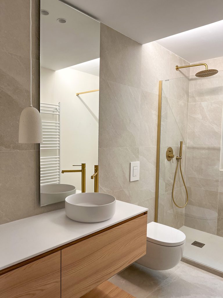 Baño diseñado por Rocío Ramírez con baldosa en tonos neutros, mueble de baño en madera y griferías en tono dorado mate. Acompaña un espejo vertical hasta el techo y una lámpara suspendida decorativa.
