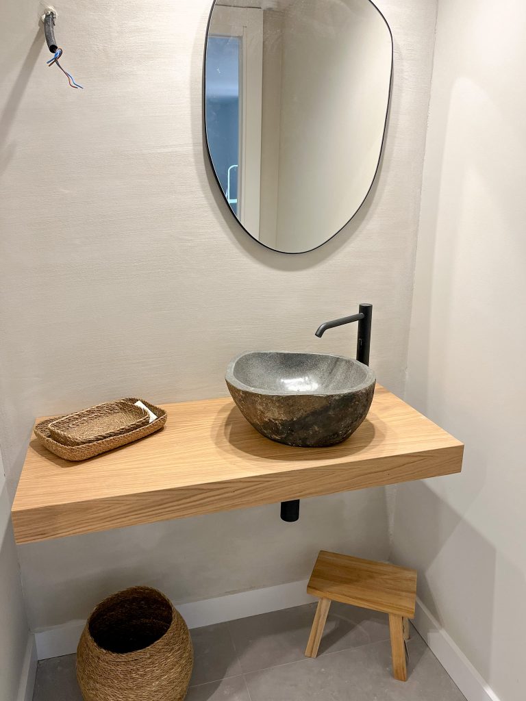 Balda de madera natural con lavabo en piedra y espejo irregular sobre pared en tratamiento de mortero de arcilla