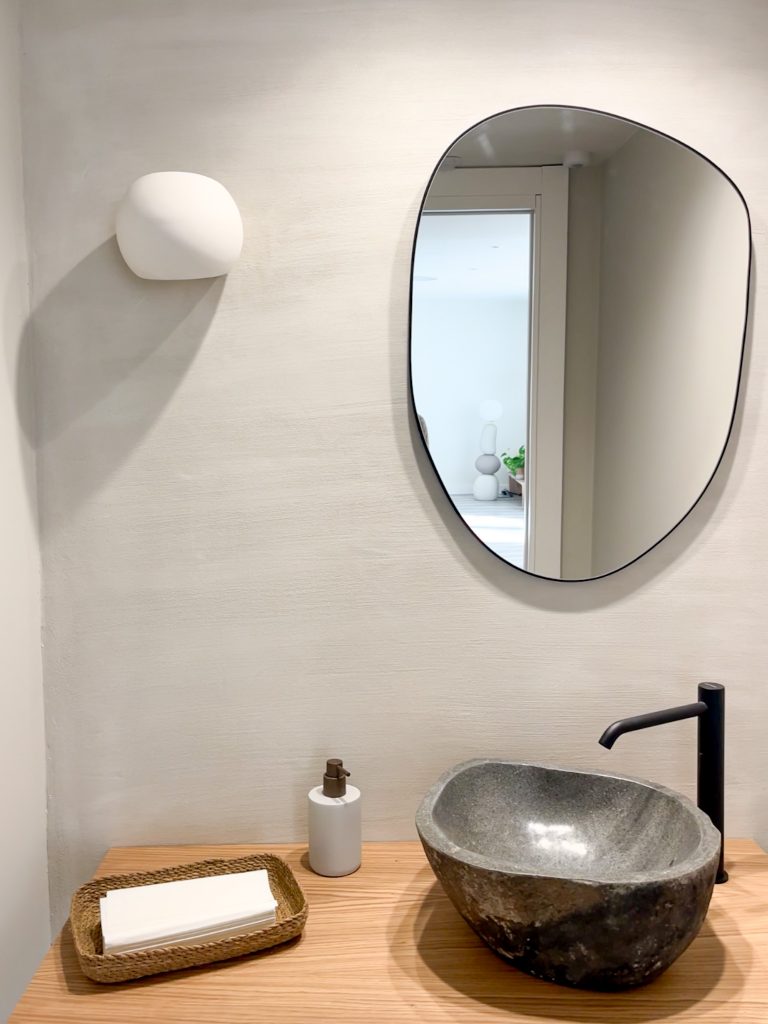 Balda de madera natural con lavabo en piedra y espejo irregular sobre pared en tratamiento de mortero de arcilla